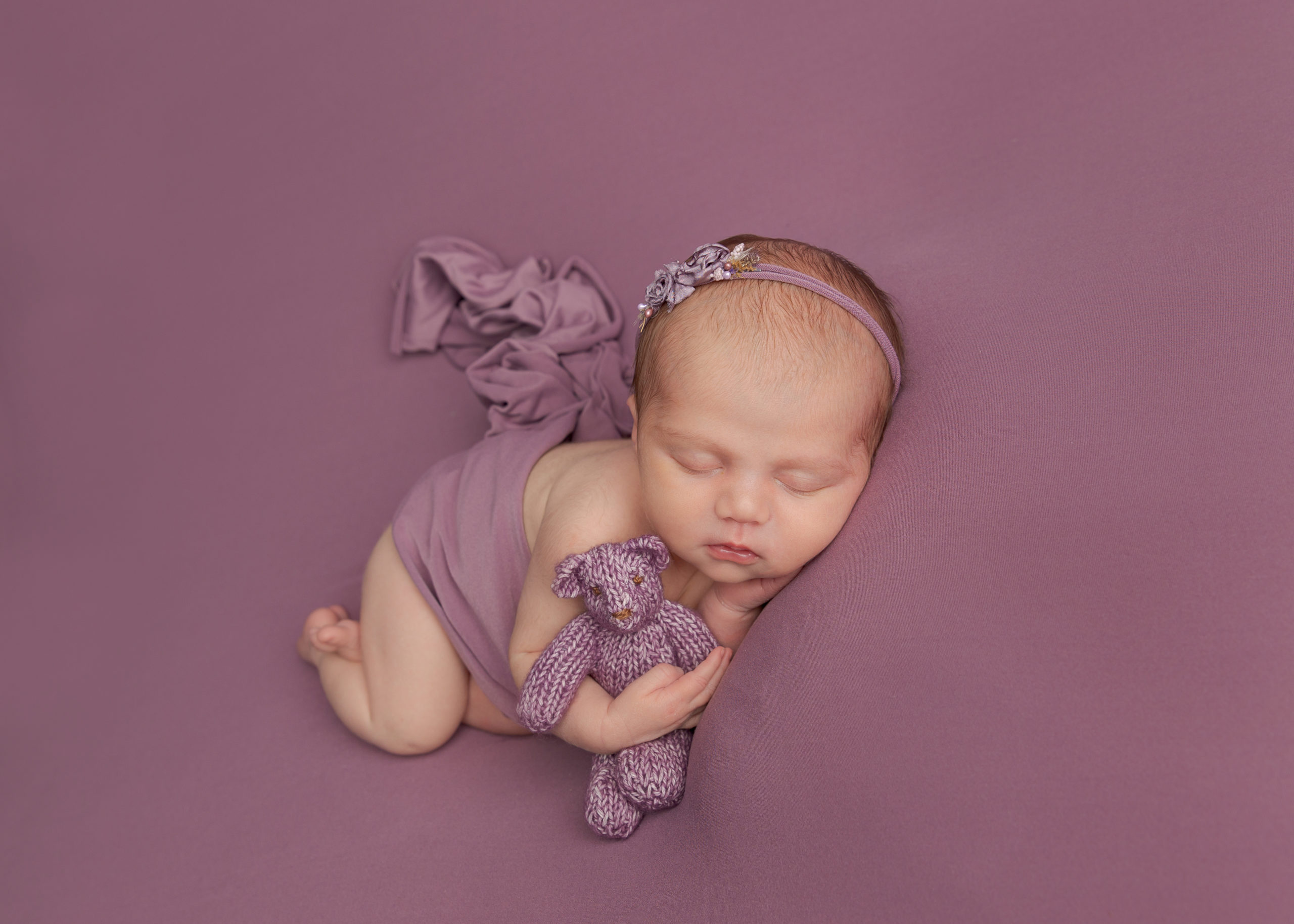 Baby girl dressed wrapped in a lilac cloth cuddling a teddy bear during a photoshoot