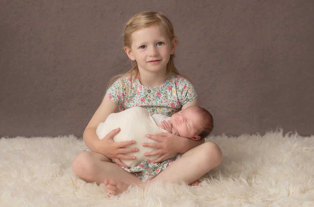 Young girl seated holding her newborn sister on her knee by Newborn Photographer Dumfries