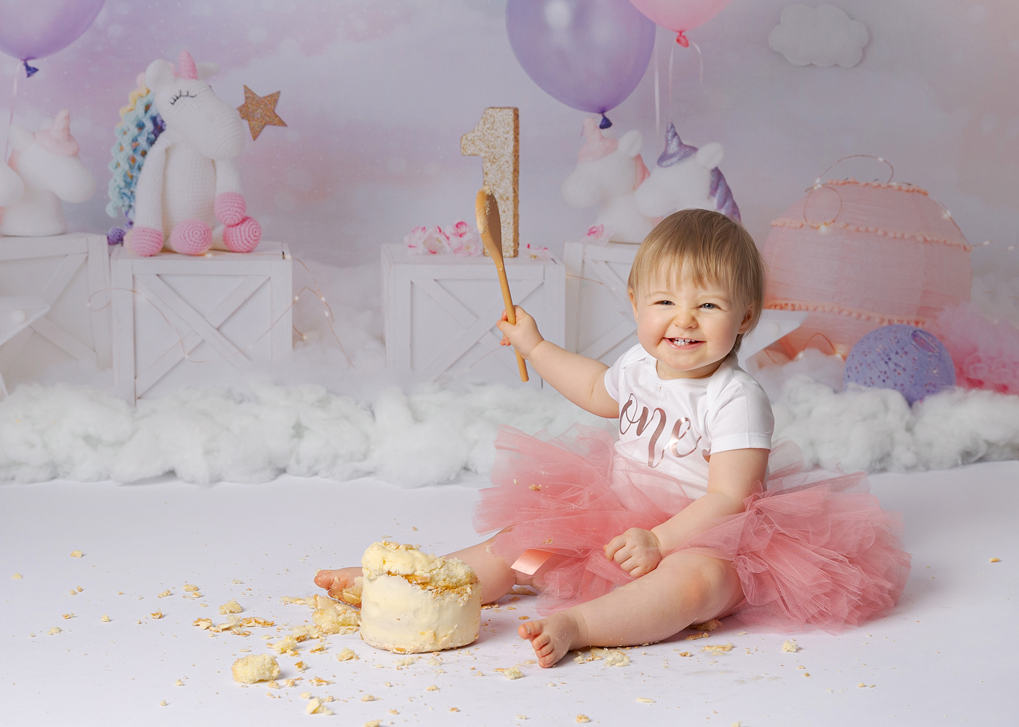 1 year old girl holding wooden spoon and smiling while smashing a cake by Newborn Photographer Dumfries