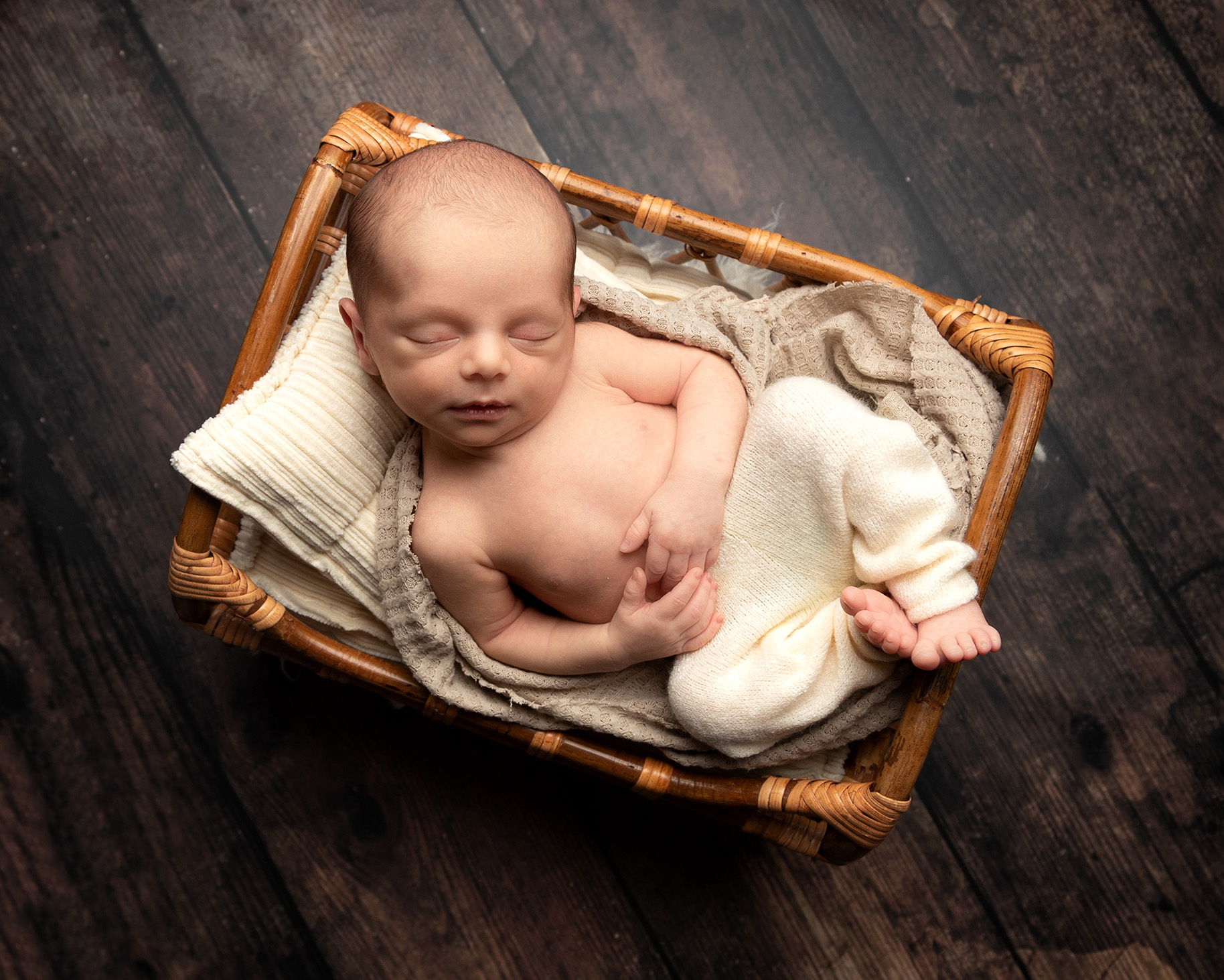 Photography of baby boy lying asleep in a small wicker bed, wearing a pair of beige trousers.  Taken during a newborn photography session by Fiona McKean Photography 